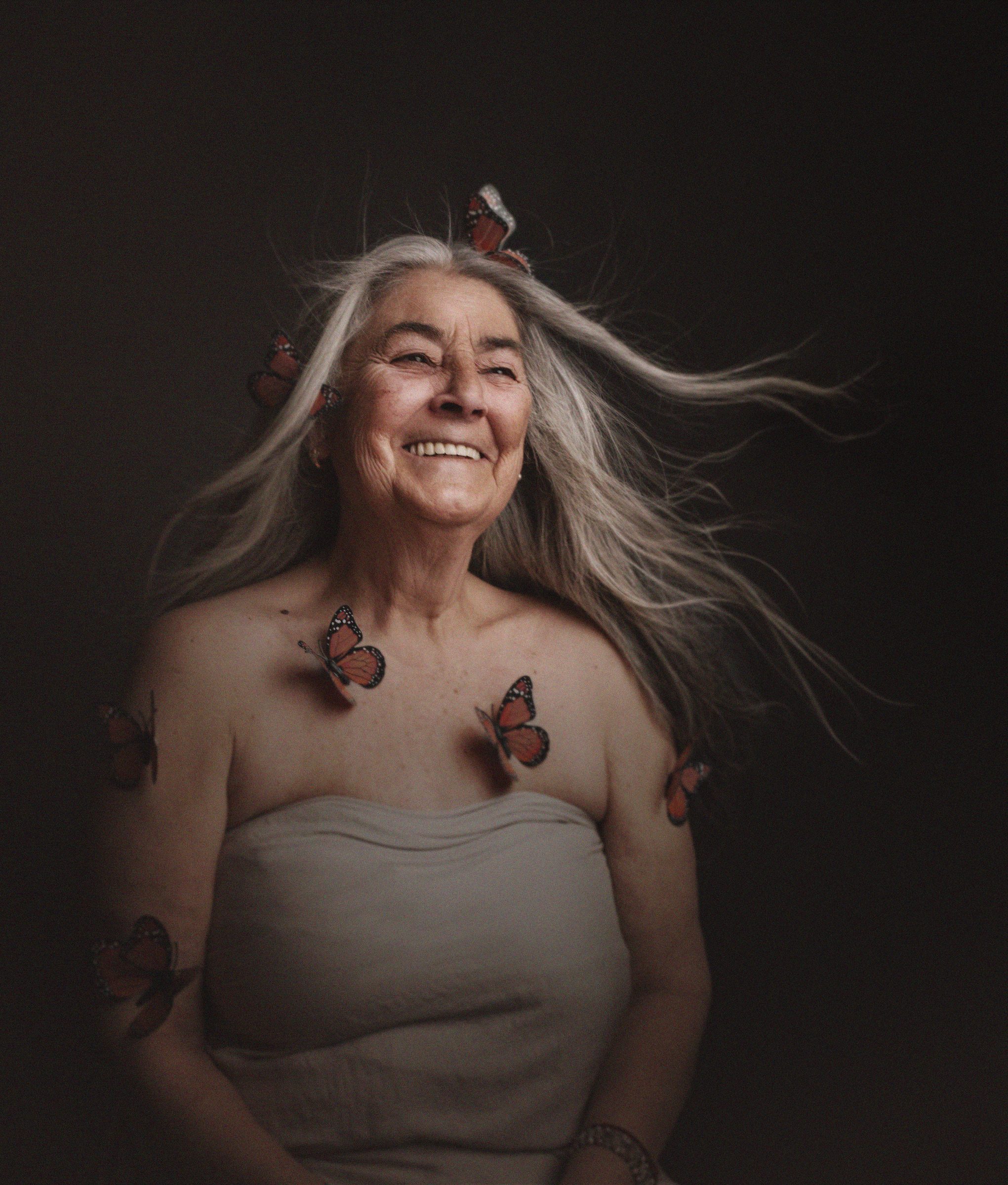 Abuela sonriendo con el pelo al viento y mariposas sobre la piel, retrato en estudio con fondo oscuro